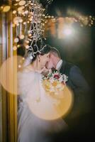A bride and groom are kissing in front of a window with lights shining through photo