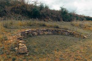 Circular stone structure surrounded by dry grass and shrubs in an open field during cloudy weather photo