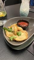 A plate of crispy, golden-brown dumplings served with a side of ketchup. The dumplings are arranged neatly on a grey plate, garnished with fresh cilantro in restaurant photo