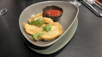 A plate of crispy, golden-brown dumplings served with a side of ketchup. The dumplings are arranged neatly on a grey plate, garnished with fresh cilantro in restaurant table photo