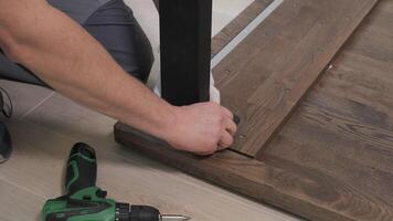 Builder in overalls screwing table legs using a screwdriver. Assembling furniture in a new apartment. video