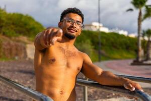 A fit young confident man with glasses and a beard indicates the camera with his hand photo