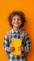 Happy boy with curly hair holding a yellow book in front of an orange background photo