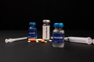 Two vaccine vials and one antibiotics vial sit next to two syringes and several pills. Health and medical concept photo