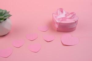 A heart shaped gift box with a pink ribbon sits next to a trail of pink paper hearts and a succulent. valentines day concept photo