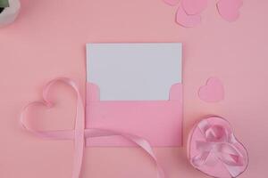 A pink envelope containing a blank card with a ribbon forming a heart shape next to a heart shaped gift box. invitation mockup photo