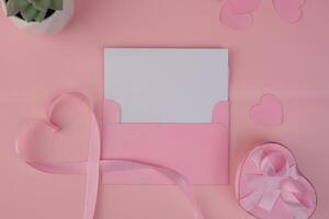 A pink envelope containing a blank card with a pink ribbon forming a heart shape next to a heart shaped gift box. invitation mockup design photo