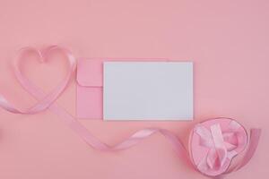 A pink ribbon forming a heart shape next to a pink envelope and a blank card with a heart shaped gift box tied with a ribbon photo