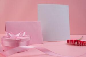 A pink gift box with a pink ribbon sits next to a pink envelope and a white card a red paper flower lies on the surface photo