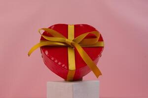 A heart shaped red gift box with a gold ribbon sits on a white cube photo