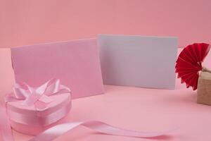 A pink heart shaped box with a pink ribbon sits next to a pink envelope and a blank card A red paper fan sits on a small wooden block photo