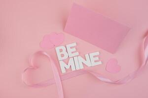 Pink hearts a ribbon forming a heart and letter blocks spelling Be Mine lay on a pink background next to a blank pink card photo
