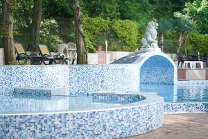 View of the hotel with swimming pool with mosaic tiles, sun loungers, a statue, a lion figure against the background of a forest, a park. Background with copy space. Concept of vacation, sunbathing photo