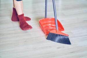 Close up view of a red broom and plastic dustpan on the floor with person's legs in red socks. Worker, cleaner using cleaning supplies with copy space. Concept of cleaning home, dust, sweeping floor photo
