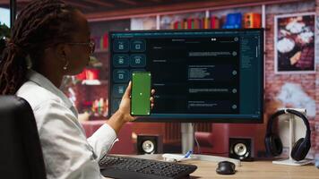 Woman seated at desk using AI LLM chatbot software and green screen phone in modern urban home office. Person uses large language model program on computer and mockup smartphone in apartment, camera A video