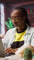 Vertical Person enjoying leisure time at home, eating snacks, texting friends on laptop. African american woman enjoying croissant, talking with mates on social network online website, using notebook, camera A video