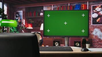 Green screen computer monitor on wooden desk in modern and stylish home workspace. Panning shot of table in home office with mockup PC display keyboard, mouse, headphones and green lamp video