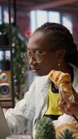 Vertical Busy freelancer in home office eating snack on the go while typing on laptop keyboard. Woman enjoying croissant while using electronic device to solve job tasks online, camera B video