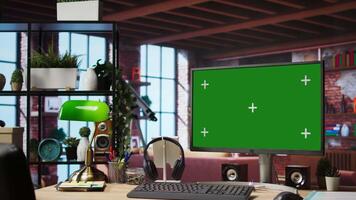 Green screen computer monitor on wooden desk in modern and stylish home workspace. Panning shot of table in home office with mockup PC display keyboard, mouse, headphones and green lamp video