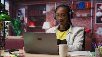 Teleworker listening music while typing on notebook keyboard in home office, writing emails. Remote employee wearing headphones using laptop, finishing project tasks, camera B video