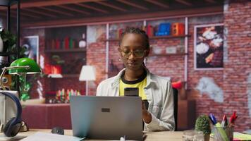 Portrait of jolly woman sitting on chair at home and texting friends on smartphone. Cheerful african american person in living room enjoying leisure time, chatting with mates, camera B video
