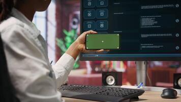 Woman seated at desk using isolated screen mobile phone to follow tutorial on AI LLM chatbot software. African american person uses mockup smartphone in apartment and GPT program on computer, camera B video