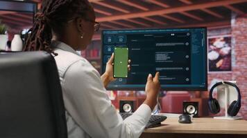 Woman seated at desk using AI LLM chatbot software and green screen phone in modern urban home office. Person uses large language model program on computer and mockup smartphone in apartment, camera B video
