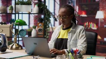Portrait of cheerful woman in home office opening laptop, starting working on tasks. Joyous african american person using electronic notebook device to do job online, camera B video