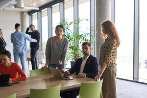 A diverse team of business professionals engaged in a discussion around a conference table in an office, while their colleagues collaborate in the background photo