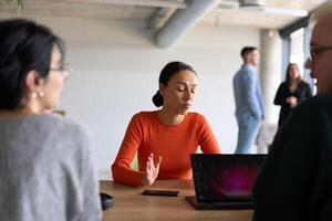 A diverse team of business professionals engaged in a discussion around a conference table in an office, while their colleagues collaborate in the background photo