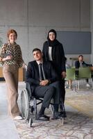 A diverse business team poses confidently, including a smiling director seated in a wheelchair, flanked by two professional female colleagues, one wearing a hijab and the other holding a tablet, in a photo