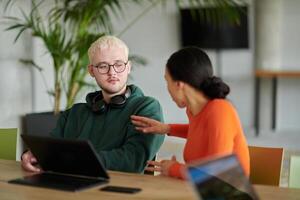 A blond businessman and his colleague sit in an office discussing work while using a laptop to collaborate on tasks photo