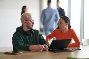 A blond businessman and his colleague sit in an office discussing work while using a laptop to collaborate on tasks photo