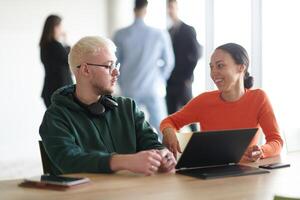 A blond businessman and his colleague sit in an office discussing work while using a laptop to collaborate on tasks photo