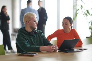 A blond businessman and his colleague sit in an office discussing work while using a laptop to collaborate on tasks photo