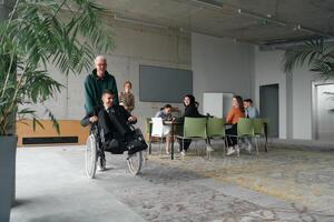 A diverse and multicultural business team collaborates in the office while their colleagues, including a director in a wheelchair and his friend, share a lighthearted moment, fostering an atmosphere photo