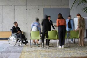 A diverse business team, working urgently around a conference table, collaborates on a meeting while their director, seated in a wheelchair, waits for their input photo