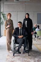 A diverse business team poses confidently, including a smiling director seated in a wheelchair, flanked by two professional female colleagues, one wearing a hijab and the other holding a tablet, in a photo