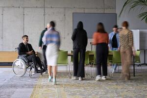 A diverse business team, working urgently around a conference table, collaborates on a meeting while their director, seated in a wheelchair, waits for their input photo
