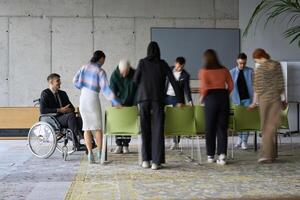A diverse business team, working urgently around a conference table, collaborates on a meeting while their director, seated in a wheelchair, waits for their input photo