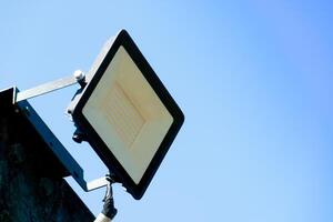 Rectangular outdoor floodlight mounted on a bracket against a clear blue sky. The light fixture is angled slightly upwards. Simple and functional. photo