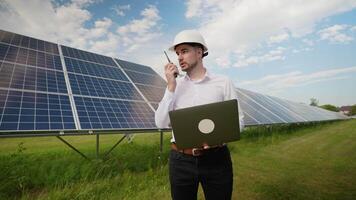 Engineer communicates via walkie-talkie while analyzing data on his laptop in a solar farm video