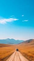 A solitary vehicle travels through vast golden grasslands under a clear blue sky in a remote mountainous region photo