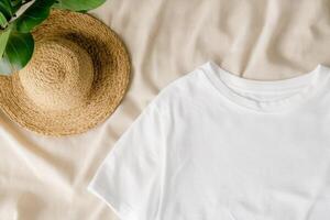 A casual white t-shirt and a straw hat placed on a soft fabric surface with green foliage photo
