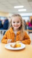 A joyful young girl in an orange jacket smiles as she enjoys her snack at a busy school cafeteria during lunchtime photo