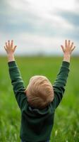 A young child joyfully raises hands towards the sky in a lush green field during a bright day photo