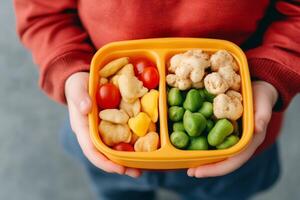 A child holds a colorful snack box filled with cherry tomatoes, crackers, and green snack puffs in a cozy indoor setting photo