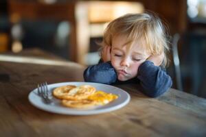 A child looking disappointed while waiting for a meal at a cozy kitchen table in the morning light with a plate of eggs in front photo
