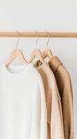 Cozy sweaters in neutral colors hanging on a wooden rack against a light wall background photo