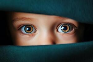 A child peers curiously from behind a fabric, showcasing striking blue eyes in a warm indoor setting during daylight photo